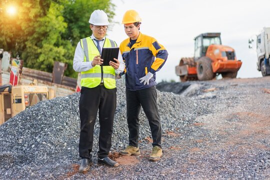 Construction Workers Inspecting Project Progress with Tablet on Site Surrounded by Heavy Machinery and Gravel
