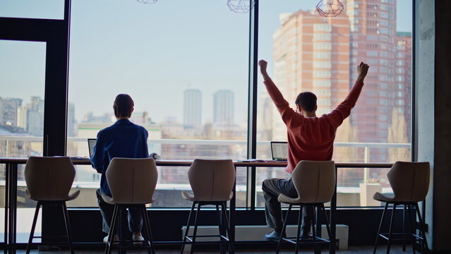 Hardworking freelancers typing laptops by cafeteria panoramic window. Tired man