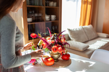 Woman making bouquet in brass vase full of dahlia flowers on dining table by cupboard. Interior and home decor. Autumn floral arrangement with pumpkins