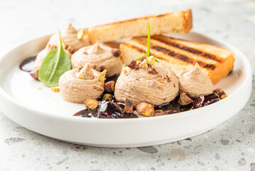 Gourmet appetizer with creamy brown mousse, chopped nuts, grilled bread, and spinach leaf on white plate. Elegant restaurant-style food presentation with dark sauce accent.