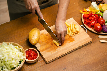 hands slicing potatoes on a wooden cutting board in the kitchen, preparing for cooking.