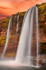 Stunning Icelandic landscape showing Seljalandsfoss waterfall illuminated by sunset, powerful stream descending against textured cliffs under colorful evening sky.