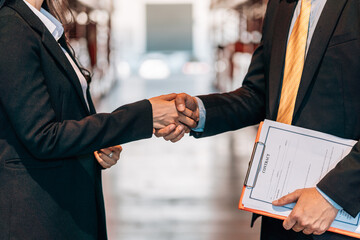 A male and female professional in business suits shake hands in a warehouse, with the man holding a clipboard.