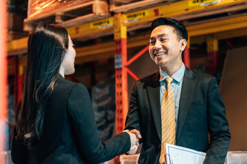 A male and female professional in business suits shake hands in a warehouse, with the man holding a...