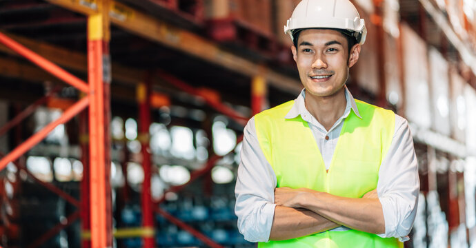 A male warehouse worker in a white hard hat and bright safety vest stands with his arms crossed, smiling slightly and looking to the side. The photo is a close-up