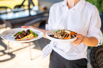 Waiter holding two plates with gourmet food: mashed potatoes with meatballs and herbs, grilled bread with mushrooms and fruit sauce. Outdoor restaurant setting, sunny patio