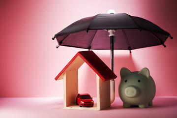 piggy bank, toy car house model under a small black umbrella against pink background