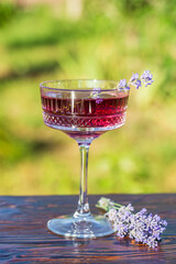 Bright cocktail or lemonade served in elegant crystal glass on wooden table against of a green summer garden. Purple color drink with lavender herb flowers, closeup