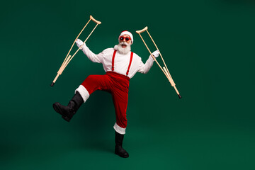Festive Santa Claus in a Stylish Pose with Wooden Crutches Against a Solid Green Background