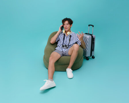 Asian man traveler sitting on bean bag talking on mobile phone with luggage camera and headphones wearing casual striped shirt and shorts relaxed vacation concept