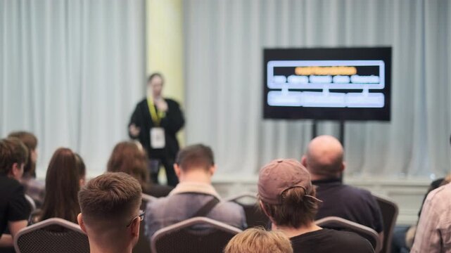 A speaker presenting to an audience in a conference setting with a monitor visible.