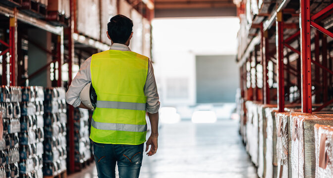 A lone male warehouse worker, seen from behind, walks down an aisle of a warehouse while holding a hard hat. He is wearing a bright safety vest.