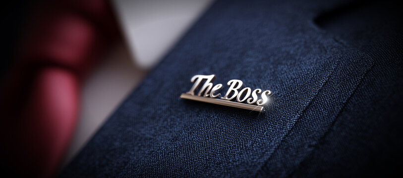 Extreme close-up of a lapel pin on a blue suit of an unrecognizable businessman with the words “The Boss” written on it in silver letters. Generative Ai.
