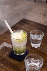 A glass of iced matcha latte with a straw sits on a wooden table alongside two glasses of water, one partially visible in the foreground