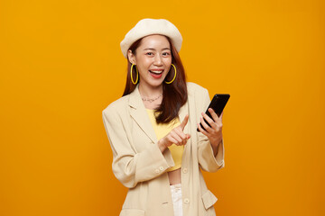 Smiling Asian woman pointing smartphone screen showing positive emotions modern digital lifestyle online communication and social media interaction isolated on yellow background