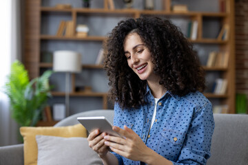 Woman with curly hair smiling and looking at a tablet, sitting on a sofa at home, enjoying online content and connectivity for entertainment and relaxation