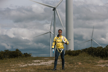 Smiling wind turbine technician in safety gear standing on green field with windmills in background. Concept of renewable energy, sustainable engineering, clean power, and green technology workforce.