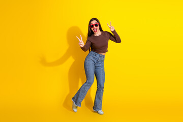 Charming young woman in casual attire showing peace signs against a vibrant yellow background with a joyful expression