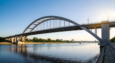 Naklejka premium A large arch bridge spans a wide, calm river under a bright blue sky. Trees line the far bank, and the sun illuminates the structure