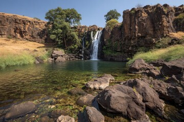 Obraz premium A tranquil waterfall cascades into a crystal-clear pool surrounded by dark, rugged rocks and lush greenery.