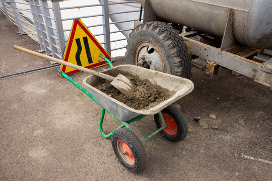 A wheelbarrow filled with concrete sits beside a warning sign and heavy equipment at a construction site. The scene depicts a typical day of manual labor and construction activity.