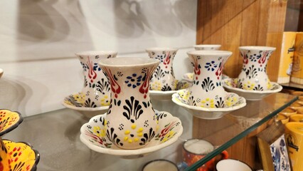Ornate Turkish tea glasses on display, showcasing traditional hand-painted designs featuring bold colors like red, blue, yellow, and black. Set on matching saucers, the glasses have a tulip shape, a c