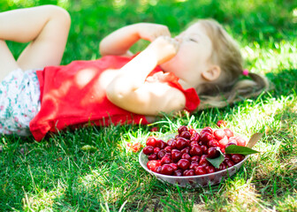 little girl in red shirt eating berries and lying on grass. a bowl with cherry on green grass in the foreground