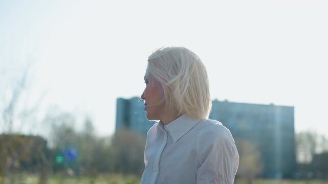 Smiling blonde woman in white shirt outdoors under bright sunlight, hair blowing in breeze, casual and relaxed expression, natural background with soft focus trees and buildings creating lighthearted