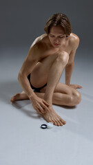 Shirtless male in black underwear applies cream to foot while sitting on floor. Concept of personal care advertising, skincare campaigns, wellness branding, and lifestyle health promotion.