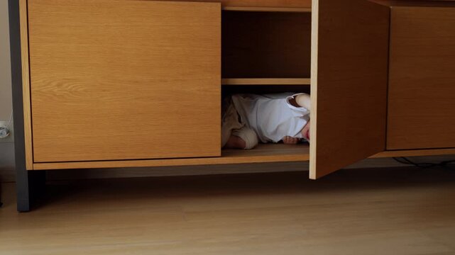 Little kid lies inside the commode hiding in it. Laughing boy opens and closes the door playing hide-and-seek.