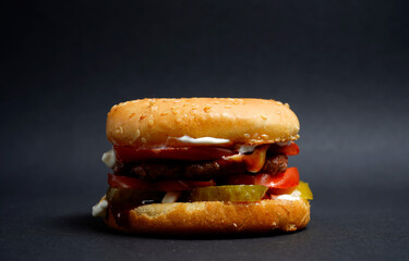 Close-up of a juicy burger with beef patty, fresh tomato slices, pickles, sauce, and sesame bun on dark background. Perfect fast food concept and street meal detail.
