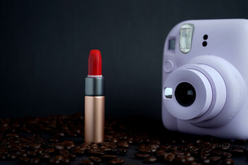 Instant white camera and red lipstick in golden tube surrounded by coffee beans on dark background....