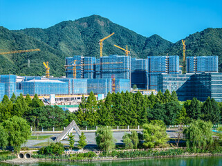 Modern glass facade commercial office buildings under construction with tower cranes against a green mountain background.