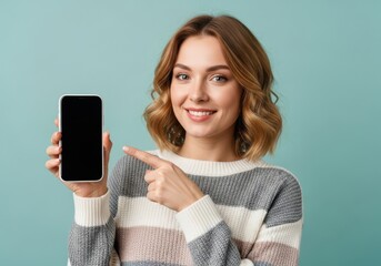 Naklejka na ściany i meble Smiling woman in a striped sweater holds a smartphone and points to its black screen, isolated against a teal background of mockup