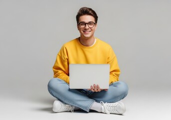 Naklejka na ściany i meble Young man with glasses wearing a yellow sweatshirt and blue jeans, sitting crosslegged with a laptop of mockup