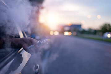 Close-up of a person holding an electronic cigarette outside a car window with smoke rising, captured at sunset on a city street.