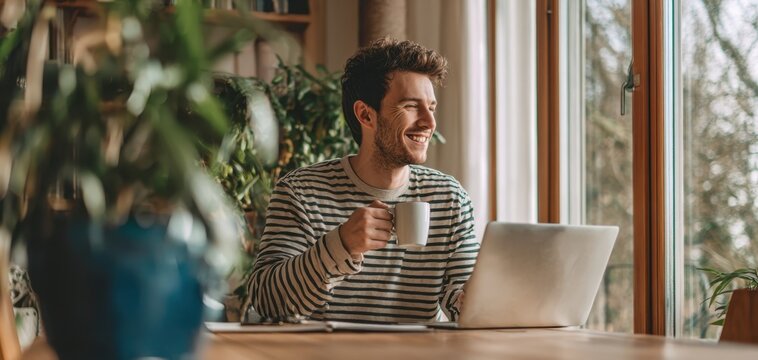 The man enjoying coffee while working on a laptop by a window at home - Powered by Adobe