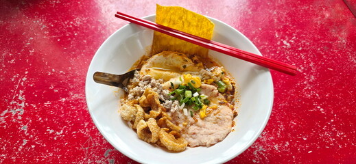 Spicy Tom Yum noodles served with fried egg, slices of minced pork, fried wontons, and pork crackling. Noodles in white bowl are placed on red table. The soup is made with chili paste and lime juice.
