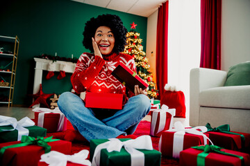 Fototapeta premium Excited woman opening Christmas presents surrounded by festive decorations at home with tree and fireplace in background