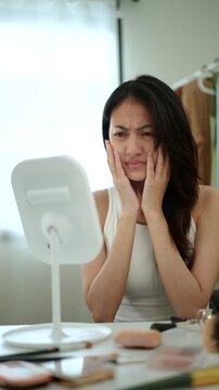 Vertical video. Worried Asian woman looking at mirror, touching dry irritated skin and acne spots. Female feeling unhappy with wrinkles and face dehydration, searching for skincare solution. 
