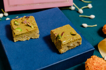 Artistic view of two square pistachio sweets, carefully placed on a blue box, surrounded by flower decorations. A colorful food photography.