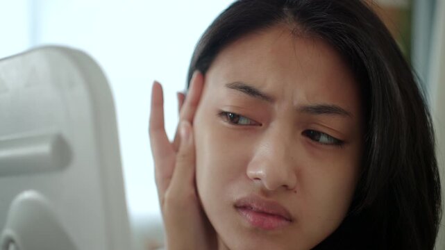 Close-up Worried Asian woman looking at mirror, touching dry irritated skin and acne spots. Female feeling unhappy with wrinkles and face dehydration, searching for skincare solution. 