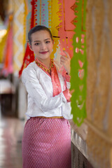 Woman in traditional Thai attire stands smiling beside colorful hanging decorations, creating vibrant and cultural atmosphere