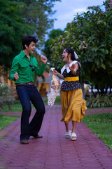 Joyful couple dances on park pathway, surrounded by lush greenery. man wears green shirt and jeans, while woman is in yellow skirt