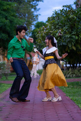 Joyful couple dances brick path park, surrounded by lush greenery. man wears green shirt and jeans, while woman is yellow skirt and white blouse