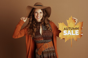 A smiling woman in brown hat and orange cardigan looks directly at camera, holding yellow maple...