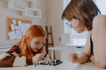 Obraz premium A teenage girl and her mother play chess in a bright living room, sharing an authentic moment of family bonding and strategic fun. Real photography capturing genuine connection