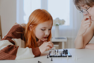A teenage girl and her mother play chess in a bright living room, sharing an authentic moment of family bonding and strategic fun. Real photography capturing genuine connection