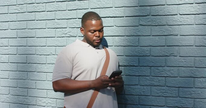 African American man with satchel leaning on brick wall scrolling smartphone smiling to camera