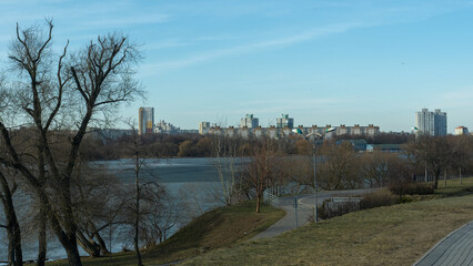 Urban waterfront park with paved walkway bare trees and street lamps overlooking midrise buildings across the lake in muted seasonal landscape. City park by the lake with walking path trees.